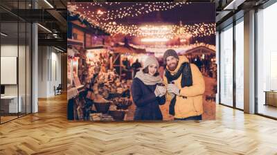 Beautiful young couple outdoors at winter on a new year`s eve with hot drinks, having fun at a winter fairy, christmas lights all around, souvenir shops Wall mural